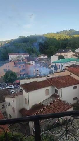 This video shows a view of a town with buildings and trees. There are multiple instances of fireworks or flares being set off, indicated by bursts of light and smoke. The fireworks appear to be red and white. The video is taken from an elevated position, possibly a balcony. The time of day appears to be late afternoon or early evening, with the sun casting a warm glow on the scene.
