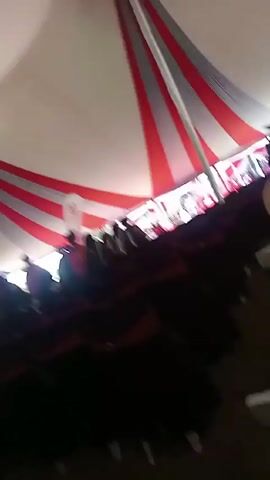 The video shows the interior of a large tent filled with people. The tent is decorated with alternating red and gray stripes radiating from the center of the ceiling. Rows of chairs, mostly black, are visible, and people wearing what appear to be graduation gowns are seated in them.  A white banner with a red logo is visible near the front. The camera moves erratically, panning across the scene. Overall, the setting suggests a graduation ceremony or similar formal event.