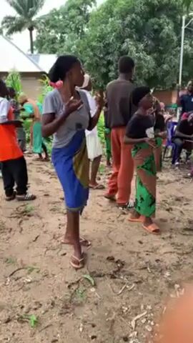 The video shows a group of people, including children and adults, dancing in an outdoor area. They are standing in a circle and moving their arms and bodies to the rhythm of the music. Some of the people are wearing colorful clothes, including traditional African prints. The background of the video includes trees, a building, and a dirt ground. The video is shot from a handheld camera and the lighting is natural.