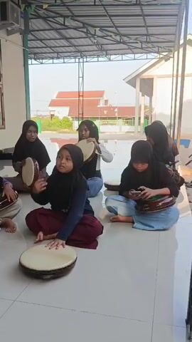 A group of young girls wearing hijabs are sitting on the floor playing drums. They appear to be practicing or performing. One girl is holding a phone while playing her drum. There are several drums visible, some are larger and some are smaller. The girls are all sitting cross-legged. The environment appears to be outdoors, under a covered patio or awning. There is a building in the background. The overall scene suggests a cultural or musical activity.