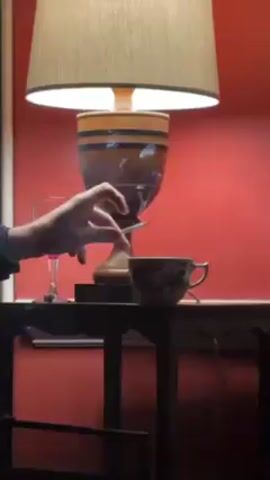 A person holding a lit cigarette is seen standing near a dark table that has a lamp, cup, and wine glass on it. The lamp is on, casting light on the objects on the table. The wall behind the table is red. Another person walks in front of the camera.