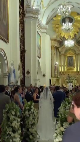 Inside a church, a wedding procession unfolds. A bride, adorned in a white gown and veil, walks down the aisle, accompanied by a man in a suit. Guests, dressed in formal attire, observe and capture the moment on their phones. The church interior features white walls, ornate gold accents, religious paintings, a statue of a saint, and grand chandeliers. Floral arrangements line the aisle, adding to the celebratory atmosphere.
