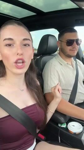 This media file captures a candid moment of two individuals, a woman and a man, inside a car. The woman is in the foreground, facing the camera with a wide, joyous smile. She has long, dark brown hair, is wearing a burgundy strapless top, a delicate necklace, and has freckles across her face and nose. She is winking and gesturing with her right hand, which has elaborately decorated fingernails. The man is in the background, driving the car. He has short dark hair, is wearing sunglasses and a light-colored collared shirt. Both individuals are wearing seatbelts.

The scene is set inside a car, with a sunroof visible above. The background suggests an outdoor setting with a road and distant landscape under a bright, possibly daytime sky. There are two beverage cups visible in the car's console. The woman's expression conveys happiness and excitement.

The overall impression is that of a cheerful road trip or outing. There is no visible text in the media file. The location context provided is Chokoba, Bulgaria.