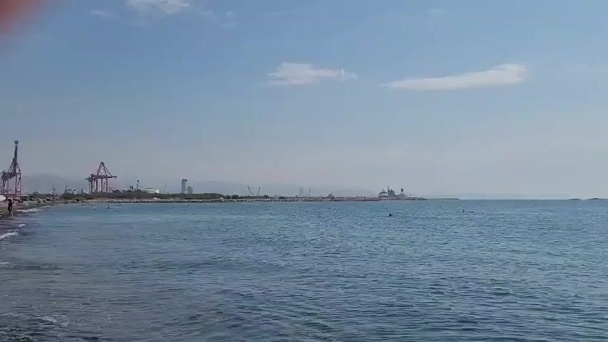 The video opens with a shot of the sea. The water is blue and calm. In the background, there is a coastline with a few buildings and some cranes. There are also a few boats in the distance. The sky is blue with a few white clouds. The video then zooms in slightly on the water, showing the gentle waves lapping against the shore. There are a few people visible on the beach in the distance, as well as some in the water. The overall impression is one of a peaceful day at the beach.