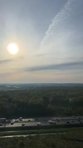 The video displays an expansive, high-angle view of a landscape featuring a multilane highway cutting through a dense green forest. Cars and trucks traverse the highway, moving in both directions. Beyond the forest, a distant skyline indicates an urban or industrial area, partially obscured by haze. The sky above is bright, with the sun visible and high in the frame. A distinct, elongated cloud formation stretches across the sky, appearing as a contrail. The overall lighting suggests daylight conditions.
