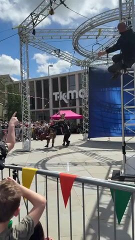 The video shows a public performance taking place in what appears to be a town square. A metal structure consisting of a circular rig suspended over two vertical ladders has been set up. Several performers in tactical gear are engaged in a choreographed action sequence. One performer is suspended upside down from the rig. Others run and perform acrobatic maneuvers below. There's a crowd of onlookers behind a metal barricade decorated with colored flags. In the background is a building identified as the "Glassworks". The sky is blue with scattered clouds, and the overall lighting suggests a sunny day. The performance is accompanied by music.