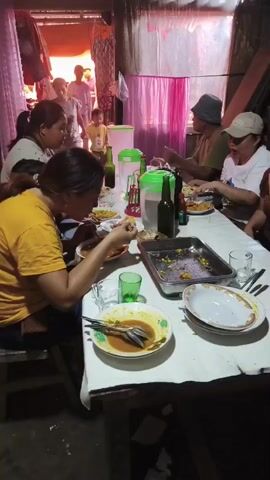 The video shows a group of people eating at a long table, covered with a white tablecloth. There are several plates of food, some half-eaten, and drinks on the table. The people are of various ages and appear to be enjoying a meal together. The environment looks like an indoor gathering, possibly a home, with simple furnishings and lighting. The overall atmosphere seems casual and convivial.