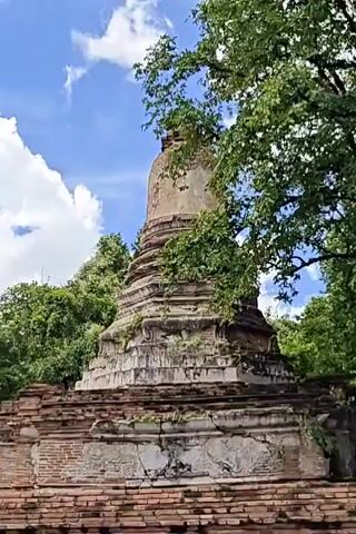 This is a short video of an ancient brick pagoda partially overgrown with vines and trees. The pagoda is made of reddish-brown bricks and has a tiered, conical shape. The sky is blue with white, puffy clouds. The video pans across the pagoda, showing it from different angles, with lush green trees surrounding it. The lighting suggests it is daytime, likely a sunny day. There are no people visible in the video, and no actions or interactions are depicted beyond the camera's movement. The overall scene conveys a sense of age and abandonment.