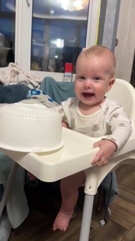 A fair-haired, light-skinned baby is sitting in a white high chair with a white salad spinner on the tray. The baby wears a white long-sleeved shirt with small bear patterns. The baby laughs loudly, then looks to its right as an adult wearing a tan shirt comes into view. The baby is inside a house with a large window behind it.