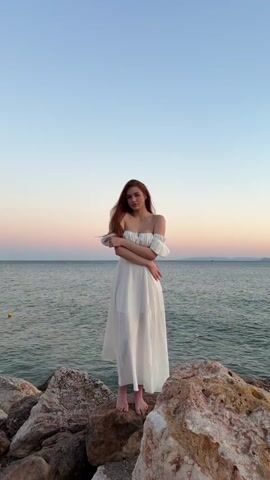 A young woman with long, auburn hair stands on a rocky shoreline, posing with her arms crossed. She is wearing a long, white, off-the-shoulder dress. The ocean stretches out behind her, and the sky above shows the soft colors of dusk. The rocks in the foreground are large and varied in color. The scene is calm and serene.