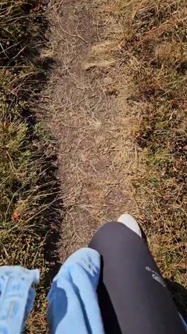 A first-person perspective video shows a person walking on a dirt path in a mountainous area. The path is lined with dry grass and shrubs. As the person walks, the view pans up to reveal a scenic landscape of rolling hills and mountains under a clear blue sky. A winding road can be seen in the distance. The video ends with a wide shot of the mountain range.