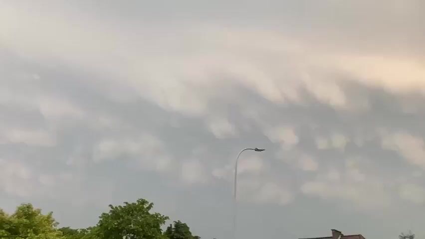 The camera pans slowly to the right, showing an overcast sky with mammatus clouds above a residential area. In the foreground, there are green trees, rooftops of houses, and a street lamp. A passing vehicle can be heard. As the camera pans, it reveals a red brick building with a “STOP” sign and what appears to be a cafe sign “Jupiter Hut.”