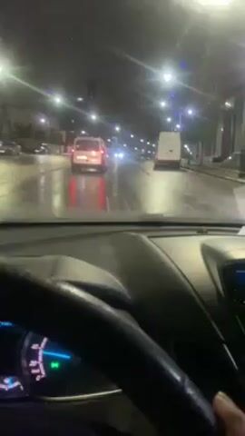 A first-person perspective video shows the dashboard of a car moving slowly along a wet city street at night. Rain falls on the windshield, obscuring the view, and the windshield wipers are in motion. Streetlights illuminate the road, reflecting in the puddles. Other cars, including a white van, are visible ahead in the same direction. The car's speedometer reads close to zero.