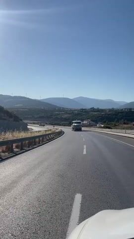 The video shows a view from a moving vehicle on a highway. A white truck is visible driving in front on the same road. The road is paved and has white lane markings. On the left side of the road, there is a metal guardrail and a rocky embankment with dry vegetation. In the background, there are rolling hills with scattered buildings and wind turbines. The sky is clear and blue with the sun shining brightly, creating a lens flare effect in the upper left corner. The overall environment appears to be a rural or semi-rural area on a sunny day.