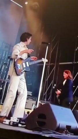 The video shows a male musician in a white suit playing an electric guitar on a dimly lit stage. He is standing and facing the left side of the frame, with his right hand on the guitar's strings and his left hand near the guitar's neck. A microphone stand is positioned in front of him. Behind him, another person is visible playing a keyboard. In the foreground, a large speaker is positioned on the stage. The background features a blurred structure with vertical lines and lighting.