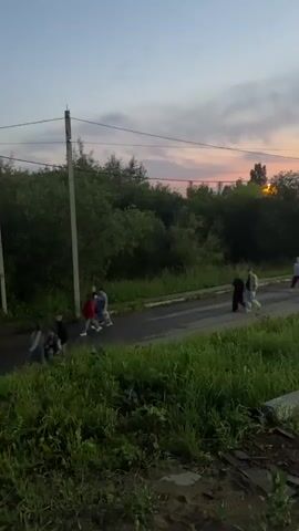The video shows a group of people walking along a paved road bordered by lush green grass and trees. The sky is a soft pastel gradient of pink and orange, indicating either sunrise or sunset. Several people are walking together in small groups, some appearing to be in conversation. One person is on a bicycle. The road appears to be a rural or suburban area, with a few buildings visible in the distance, including a building with illuminated signage. A utility pole stands on the left side of the road. The lighting suggests it is dusk or dawn, creating a serene atmosphere. The camera pans from left to right, capturing the progression of the people along the road and the changing perspective of the environment.