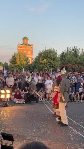 A group of people, some dressed in historical costumes resembling medieval soldiers, are gathered outdoors. In the foreground, two individuals in these costumes are facing each other, seemingly participating in a performance or reenactment. One is wearing a conical helmet, chainmail, and dark pants, and is holding a wooden sword. The other, also wearing a helmet and chainmail, is standing next to a person in a red dress with traditional embroidery and a headdress. A crowd of onlookers, dressed in modern clothing, watches the event. The background shows a tall, orange building with a green dome, trees, and a clear sky. There are fallen leaves or petals on the ground in front of the performers. The scene appears to be a public gathering or festival.