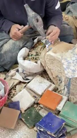 A person is seated cross-legged on the ground, surrounded by piles of colorful ceramic tiles and fragments. They are using a hammer and a small chisel-like tool to break a small piece of a white tile into smaller fragments. These fragments are being added to a bag already filled with similarly sized pieces. The person is wearing dark clothing. The setting appears to be a workshop or studio. The environment is dusty and cluttered.
