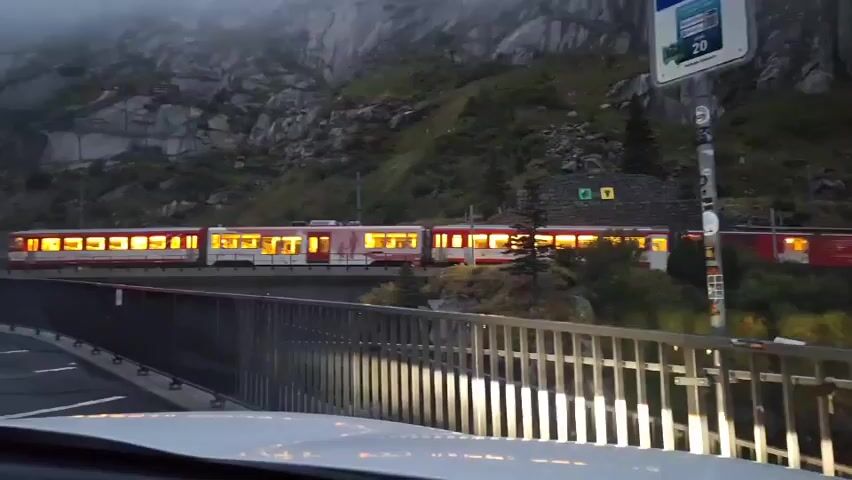 A red and white train travels on a track in a mountainous area. The train has lit windows, suggesting it is occupied. The train is moving from left to right. A car's headlights illuminate the foreground, indicating the viewer is inside a vehicle. A metal railing is visible in front of the car. A sign with a blue parking symbol and numbers is mounted on a pole to the right of the train. The environment is rocky and appears to be a mountain pass, possibly with fog or mist in the background. The time of day appears to be dusk or early morning due to the lighting conditions.