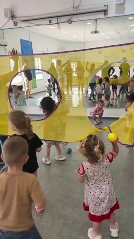 In an indoor activity room, likely a kindergarten or daycare, children are actively engaged with a large, colorful parachute. The parachute is made of yellow fabric with purple trim and has several circular openings. Some children are under the parachute, appearing to be playing with small, colorful balls. Others are standing around the edges, interacting with those inside or observing the activity.

In the foreground, two young girls and a boy are visible. One girl in a black shirt and pink shorts looks towards the parachute. Another girl, in a pink and white patterned dress, is holding a yellow ball and appears to be participating in the game. The boy in the foreground is wearing a tan t-shirt and denim shorts and is looking towards the parachute.

In the background, through the transparent parachute, a group of adults and children can be seen. The room has a mirrored wall, reflecting the scene and giving the impression of a larger space. The lighting suggests it is daytime, and there are no visible cues about the weather. The overall atmosphere appears lively and energetic, with children likely experiencing joy and excitement during this interactive play session.

The location is identified as Kiryat Bialik, Israel. No visible text is present in the image.