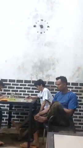 Three people are sitting at a table. One person is eating, another is looking down at the table, and the third is in a chair with his legs crossed. There is a clock on the wall. The wall is white and has a black brick pattern on the bottom half.