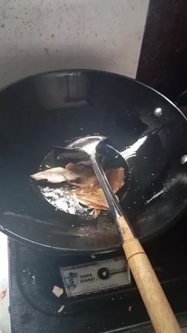 A close-up, overhead shot shows fish being fried in a black wok on a gas stove. The wok is dark, and some oil is visible with the fish submerged in it. A metal spatula with a wooden handle is being used to flip the fish. The gas stove is black and has a dial with the word "TANPA SYARAT" visible on it. The background is a light-colored wall. The video is short and has no sound.