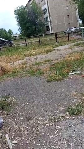 The video captures a ground-level view of an outdoor area with dry grass, dirt, and scattered debris. In the foreground, a person's legs are visible, clad in patterned pants, sitting on what appears to be a bench. Next to them is a plastic bag filled with various items. A discarded tire, painted green and blue, is visible in the midground, amidst overgrown grass. Beyond this, a black car is parked near a fence. In the background, a multi-story building stands, with trees and more parked cars in front of it. The sky is overcast. The camera pans slightly, showing a playground structure with an archway in the distance.