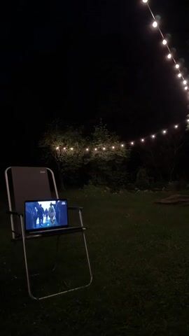 The video captures an outdoor scene at night. A folding chair is positioned on a grassy lawn. A laptop is open and placed on the chair's tray, displaying a video of a group of people. String lights with individual bulbs are visible in the background, illuminating the trees and dark sky. The overall ambiance is serene and dimly lit.