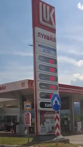 A gas station with red flags and a white canopy is visible against a bright blue sky with some clouds. Trees and foliage line the bottom of the frame, suggesting a roadside view. The video appears to be captured while moving, with the camera panning across the scene.