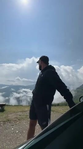 The video shows a man in athletic wear, including a black hoodie and shorts, standing next to an open car door. He is wearing a baseball cap and sunglasses. The man is outdoors, with a scenic view of mountains and clouds behind him, suggesting he is in a hilly or mountainous region. The sun is shining brightly in the sky. The man is seen looking away from the camera and then turns to face it. He appears to be leaning on the car.