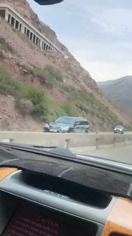 The video shows a car driving on a mountain road. The road is paved and has a concrete barrier separating it from the side of the mountain. The mountain is covered in rocks and sparse vegetation. There is a tunnel or bridge structure built into the side of the mountain, with arched openings. Another car is visible driving in the opposite direction. The interior of a car is visible in the foreground, showing the dashboard and part of the steering wheel.