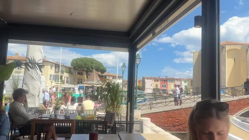 The video shows a young woman sitting at a table in an outdoor restaurant.  She has blonde hair and is wearing sunglasses on her head.  The restaurant has a view of a canal with boats and buildings on the other side.  There are other people in the restaurant, and people walking on a bridge over the canal.  The sky is blue with white clouds.  The camera pans over to show the boats in the canal, including a covered boat right next to the restaurant and other larger boats further away.
