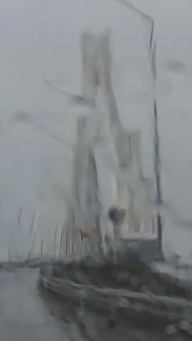 The video captures a dynamic perspective from inside a vehicle driving across a large cable-stayed bridge during a heavy rain shower. The primary subjects are the bridge itself, including its towering white pylons and numerous supporting cables, and the wet road surface.

The weather is a dominant element, characterized by a gloomy, overcast sky and persistent, heavy rain. Streaks of water are visible on the windshield, indicating continuous rainfall and slightly obscuring the view. Visibility is somewhat reduced due to the precipitation.

The bridge's architecture is prominent, featuring two tall, sleek, light-colored pylons that angle inward, from which a multitude of cables extend down to the bridge deck. These cables create a striking V-shaped pattern. Streetlights line both sides of the road, and a concrete guardrail runs along the right edge. On the right side of the road, a small, utilitarian structure, possibly a control room or utility building, is visible next to a pole. A yellow sign is partially visible in the background, but its text is unreadable due to the distance and weather.

The activity depicted is the act of driving across this impressive structure. The camera's movement suggests the vehicle is in motion, navigating the wet conditions. The time of day appears to be daytime, but the heavy cloud cover and rain create a somber, grey atmosphere. The scene is set in what is identified as Im. Voykova, Russia, likely depicting the Volgograd Bridge. No specific people are visible, but the presence of the vehicle implies an occupant.