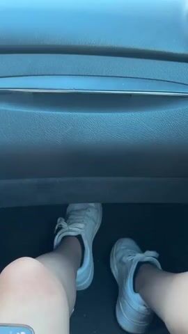The video begins with a low-angle, interior shot of a car's dashboard and the feet of the driver wearing white sneakers. The perspective then shifts to the windshield view as the car is in motion. The scene outside the car reveals a rural landscape with green fields, trees, and rolling hills under a sky with scattered clouds tinged with pink and orange, suggesting it is either sunrise or sunset. The camera continues to focus on the road ahead, which winds through the scenic countryside.