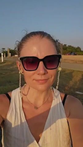 A woman with blonde hair pulled back and sunglasses on smiles at the camera. She wears a white top and a gold necklace. A beaded strap hangs from her sunglasses. She is outdoors in a grassy area with trees and a clear sky.