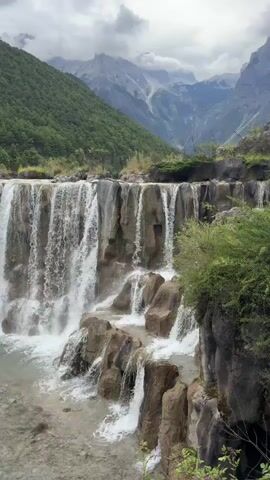 A scenic view of a tiered waterfall cascades over rocks with lush green mountains in the background. The water flows down into a clear stream. The sky is overcast with clouds.