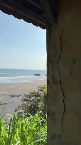 The video shows a tropical beach with lush green vegetation in the foreground. A weathered stone wall with a large crack runs vertically along the right side of the frame. Beyond the wall, a sandy beach stretches towards the ocean. Several rock formations rise out of the sea. Waves are crashing against the shore. In the distance, a few people can be seen walking on the beach. The sky is clear and blue. The camera pans slightly to the right, revealing more of the beach and coastline.