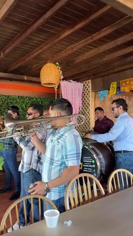 A band is playing music indoors, likely at a restaurant or venue with a thatched roof and decorative pink drapes. In the foreground, a man in a blue plaid shirt plays a trombone. Behind him, another man in a plaid shirt and sunglasses also plays a trombone. To the right, a man in a white and blue plaid shirt plays drums. Another man in a plaid shirt stands behind him. The band is performing for an unseen audience.