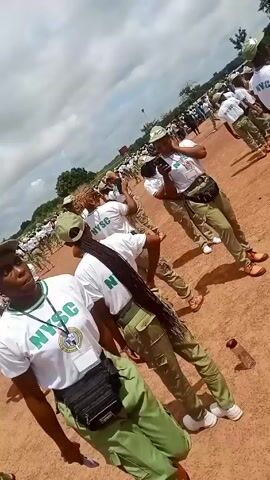 The video shows a large group of people outdoors, dressed in similar white T-shirts with the acronym "NYSC" in green, paired with khaki pants and hats, all standing on reddish-brown ground. Many are holding or adjusting their hats, and some have bags around their waists. In the background, more people in the same attire can be seen. The weather appears cloudy, with a few trees visible.