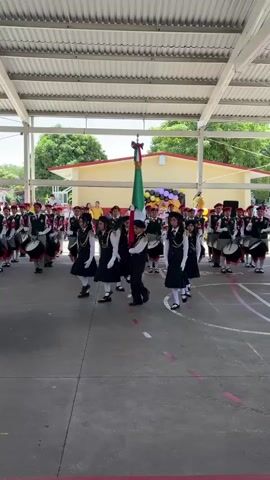 The video shows a school marching band parade. A group of students dressed in matching uniforms, a mix of dark skirts and light tops for girls and dark pants and light shirts for boys, are lined up in formation. The girls are wearing white sashes or scarves. Some of the students are carrying drums and appear to be playing them. In the foreground, a few girls are dancing or performing a coordinated movement. A large Mexican flag is being held upright on a pole, positioned in the center of the formation. In the background, there are buildings that appear to be part of the school, with a covered walkway or awning. There are also some decorations, including balloons. The ground is paved, possibly a schoolyard or courtyard. The overall atmosphere seems to be one of a celebratory school event.
