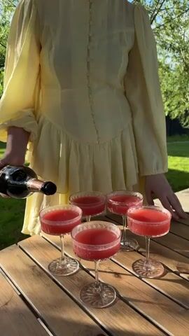 A woman wearing a yellow, long-sleeved dress is pouring a dark bottled beverage into five stemmed glasses filled with a red liquid. The glasses are arranged on a wooden picnic table outdoors, with green grass and trees visible in the background. The focus is on the glasses and the action of pouring the drink.