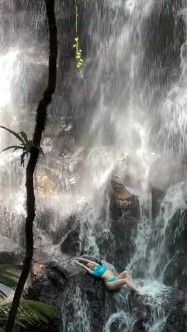 The video displays a picturesque natural setting featuring a powerful waterfall in Gianyar, Indonesia. A young woman, dressed in a blue and light green two-piece swimsuit, is prominently featured, lying on a large, dark rock formation directly in front of the cascading water. Her pose is relaxed, with her arms extended slightly above her head, and she appears to be looking upwards, enjoying the scene.

The waterfall is the dominant feature, with water rushing down a rugged, moss-covered rock face, creating a misty atmosphere. The surroundings are rich with tropical foliage, including dense green plants and a large, moss-covered tree trunk with vines on the left side of the frame, indicating a lush jungle or rainforest environment. The lighting suggests daytime, with bright light filtering through the falling water, creating a luminous effect. No visible text appears in the video. The audio accompanying the visuals is the immersive sound of the rushing waterfall. The overall mood conveyed is one of tranquility and natural beauty.