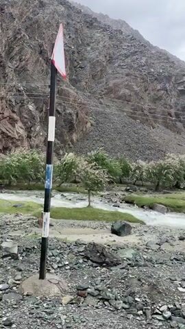 The video shows a POV shot from a vehicle traveling along a rough, rocky road. The road is bordered by a fast-flowing, rocky river on the left and steep, barren, rocky mountains on the right. Numerous small, green trees with white flowers line the bank of the river, creating a stark contrast with the surrounding arid landscape. Power lines are visible running along the side of the mountain. The sky is overcast and cloudy, suggesting a cool or possibly rainy day. The vehicle's rearview mirror is visible at the bottom left of the frame in the first few seconds. The scene conveys a sense of remote, natural beauty and the ruggedness of the mountainous terrain.