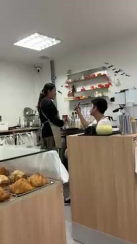 The video shows a brightly lit bakery café decorated for Halloween. A woman in a black shirt and tan apron stands behind the counter, speaking to a young man who is making a drink. The man glances at the camera and smiles. Pumpkins, ghosts made of white sheets, and paper bats adorn the counter area. The café also has a pastry display case filled with croissants. The camera then pans to a small table where two women are sitting, enjoying drinks and pastries.