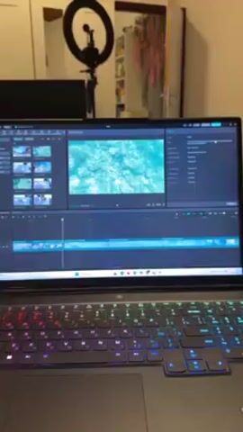 A woman is editing a video on a laptop. The laptop is on a bed. The laptop has a colorful keyboard. There is a white microphone with a grey windscreen on the bed next to the laptop. The woman has pink and purple hair. She is wearing headphones. The video being edited shows a blue watery pattern. The room has a ring light, a television, and decorations. The woman says "I'm still editing this video, talking with fans, and".
