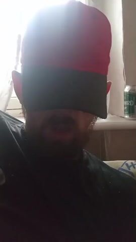 A person wearing a red baseball cap and a black blindfold is exhaling a cloud of white vapor. The person has a beard and appears to be wearing a black shirt. The background shows a window with white curtains and a can of what appears to be a beverage on a windowsill. The person is seated, and the scene is lit from the left.