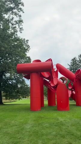 A large, red abstract sculpture is centered in a grassy park under a cloudy sky. Trees are visible in the background. The sculpture is composed of large, hollow cylindrical and tubular shapes connected in a seemingly chaotic yet balanced arrangement. Some of the tubes are wide and open, while others are narrower and appear to be attached to larger vertical supports. The sculpture has a playful, almost organic feel despite its industrial material and bold color. The grass is well-maintained and covers the ground surrounding the artwork. The lighting is diffused due to the overcast sky, casting soft shadows. There are no people visible in the frame.