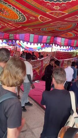 The video shows an inside view of a large tent filled with people browsing through clothing racks. The tent's ceiling is vibrantly decorated with red, orange, and green patterns. The crowd mainly comprises adults of varying ages and ethnicities. Some individuals are examining clothing hanging on racks, while others are in conversation or moving through the space. The clothing on display appears to be a mix of formal and casual wear. The floor inside the tent is covered with a red carpet. The overall atmosphere seems like a busy market or clothing sale event.