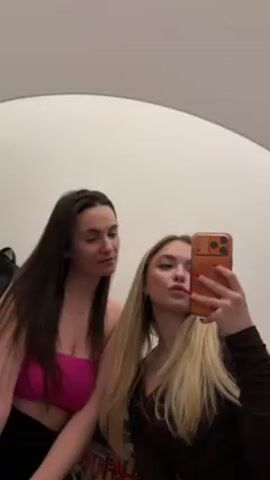 Two young women are taking a selfie in a mirror. One woman has long, dark hair and is wearing a hot pink strapless top and black pants. The other woman has long, blonde hair and is wearing a dark brown top. She is holding an orange phone and making a kissing face. The woman with dark hair is smiling. The background is a plain white wall with a partial view of clothing and a colorful item.