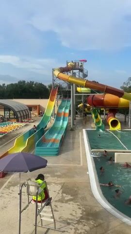 The video shows a water park on a sunny day. There are several large water slides, one multi-colored tube slide and two open slides with a green and orange design. People are swimming in a pool at the base of the slides. There are several lounge chairs set up around the pool. A lifeguard is seated in a lifeguard chair. The video pans from left to right, showing more of the pool area and then the surrounding trees and mobile homes.